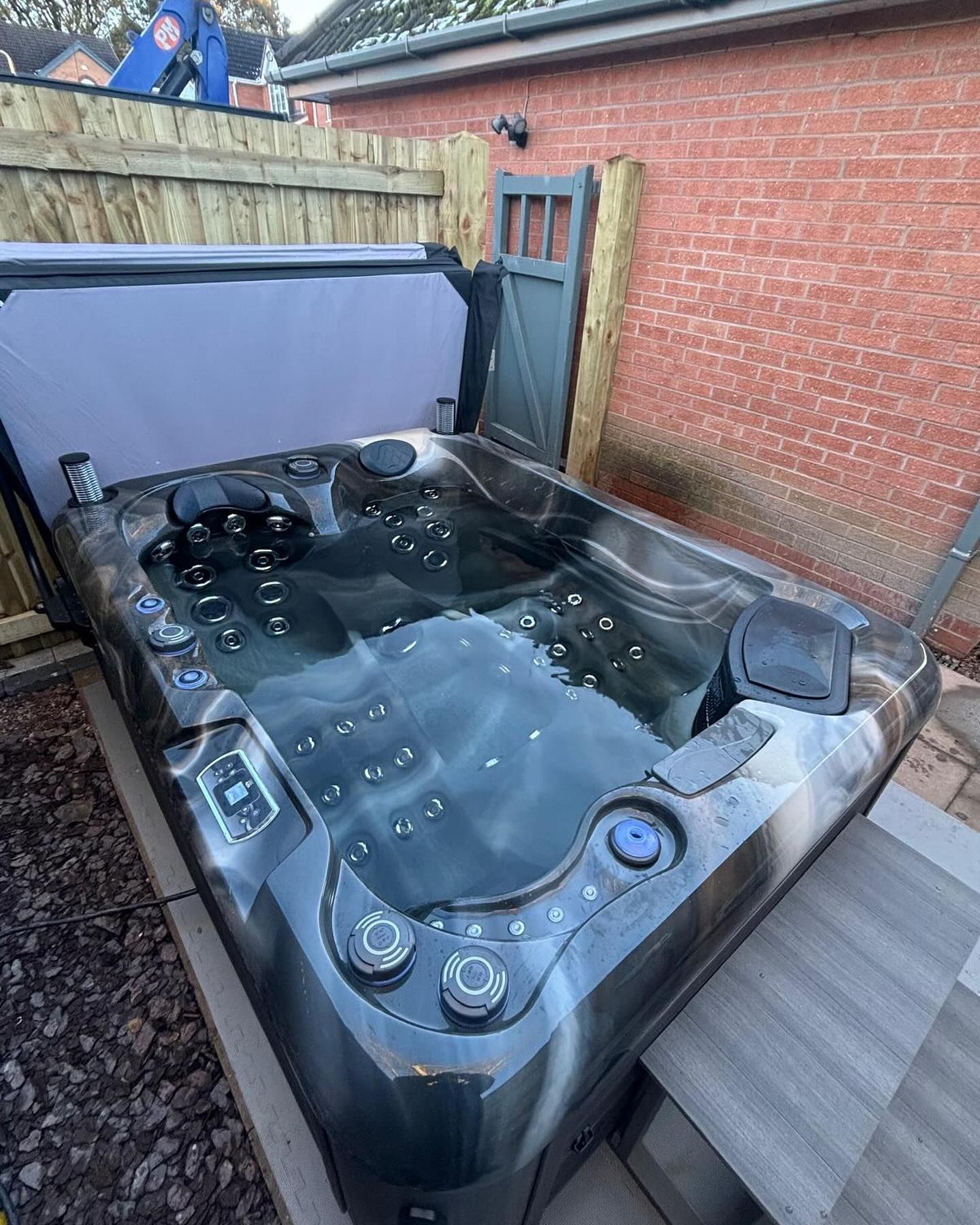 Dark hot tub with water, open cover, controls, surrounded by brick wall and wooden fence.