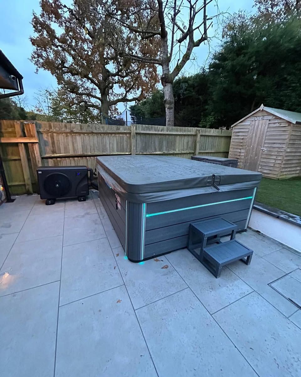 Hot tub with gray siding and cover on a stone patio with a heat pump, steps, and a wooden fence.