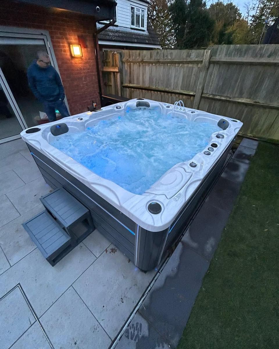 Hot tub in backyard with steps; partially visible person. Water illuminated, gray surround and steps, green grass.