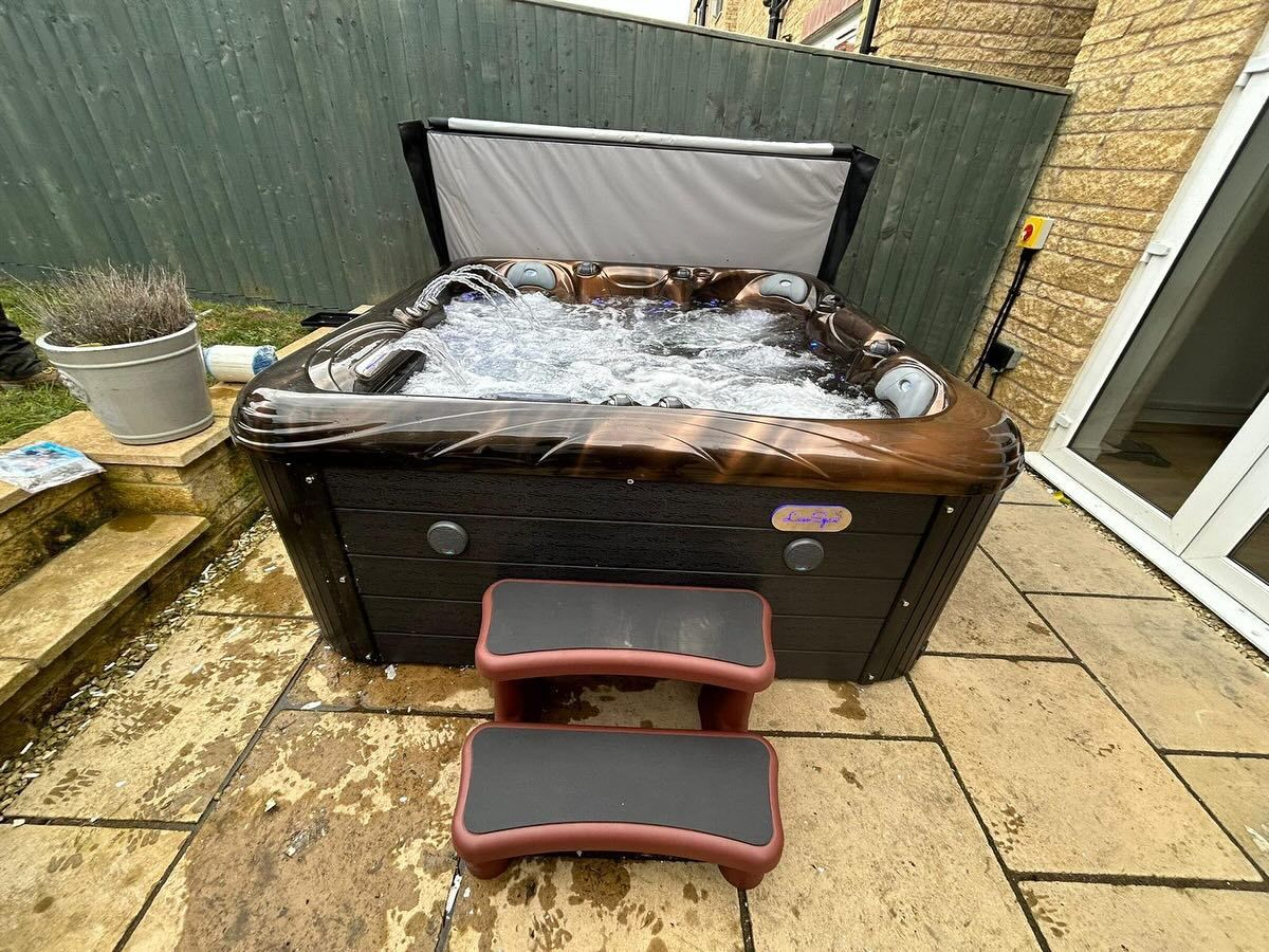 A hot tub with bubbly water, brown exterior, and red steps, on a stone patio in a backyard.