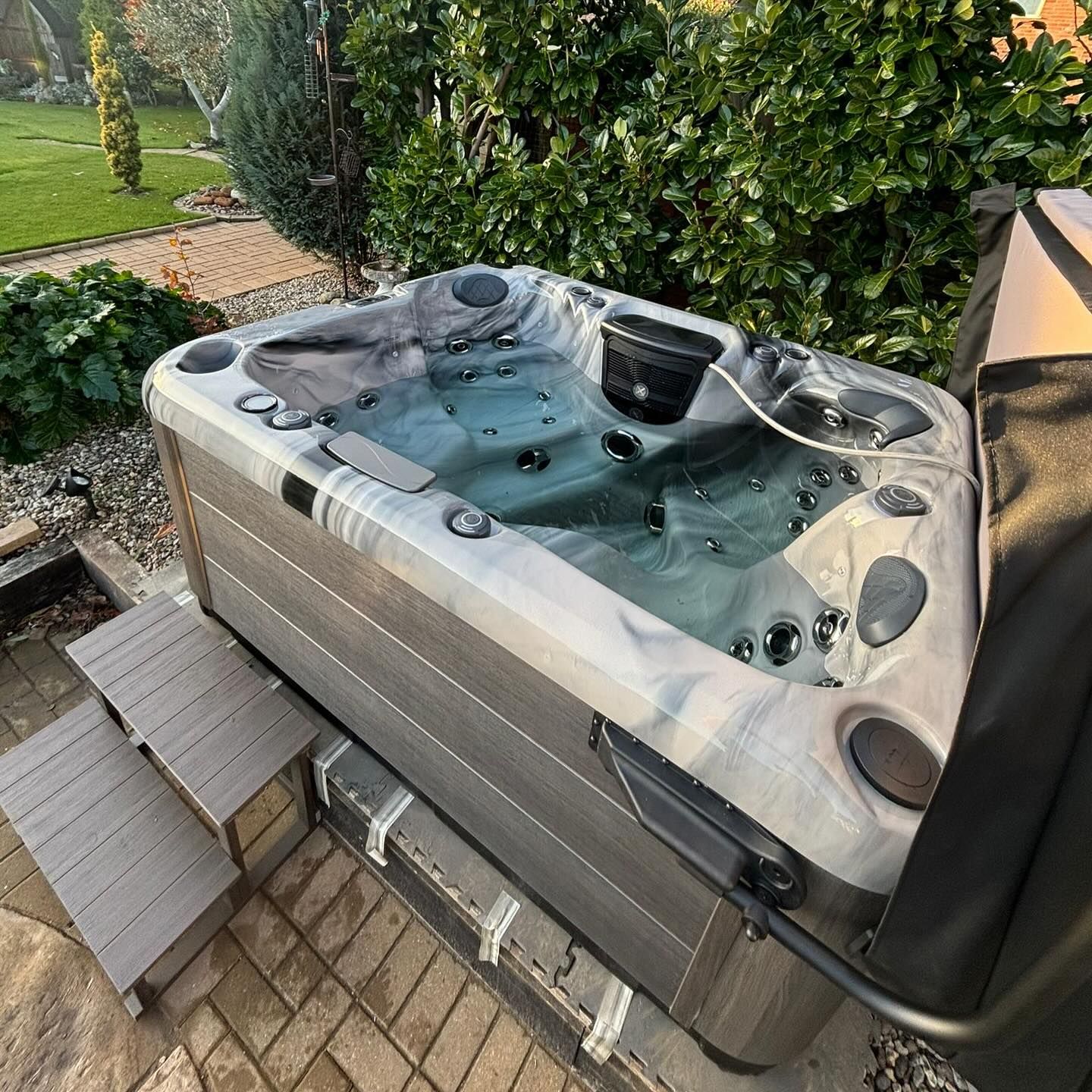 Hot tub in a backyard setting, surrounded by greenery. Gray steps lead up to the tub's edge.