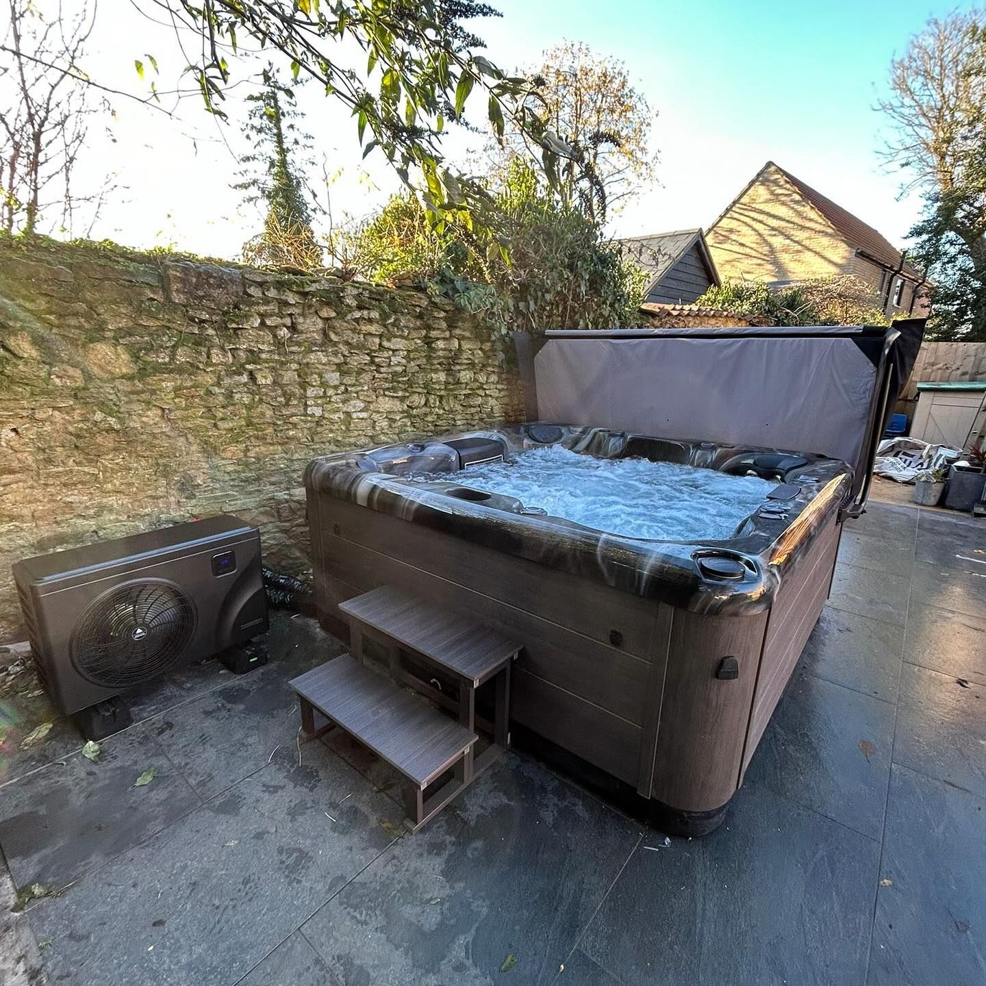 Hot tub on a patio with steps, open cover, stone wall in background.