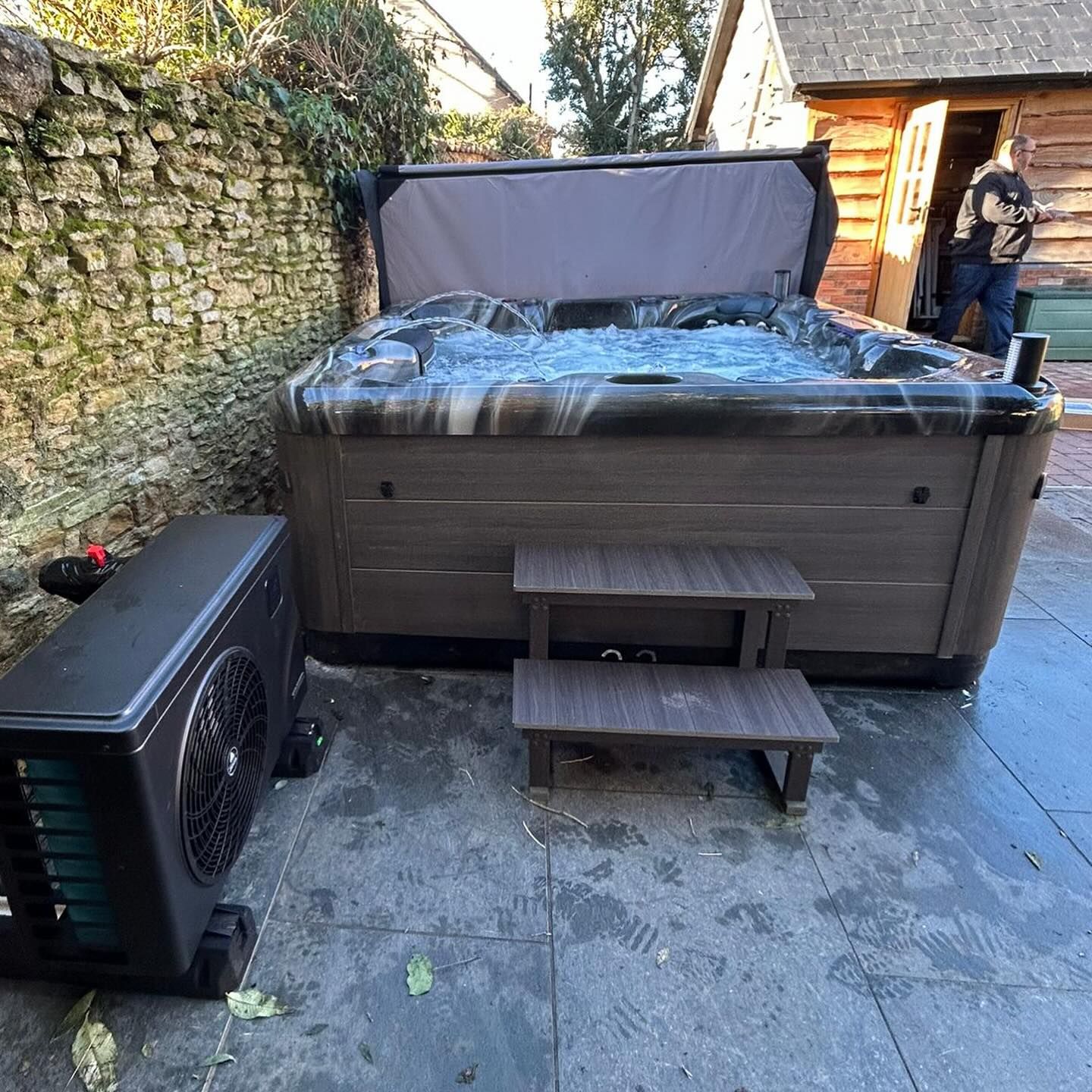 Hot tub with open cover, steps, and attached heater, outside on stone patio.