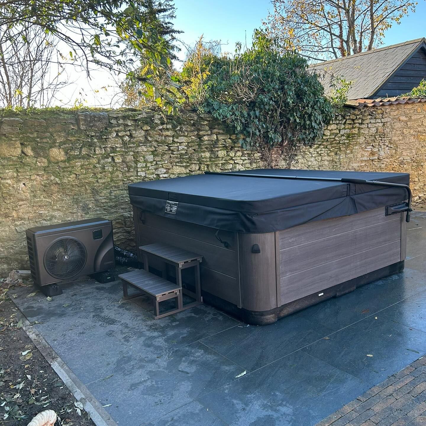 Hot tub with cover on a patio, next to a brick wall and heat pump.