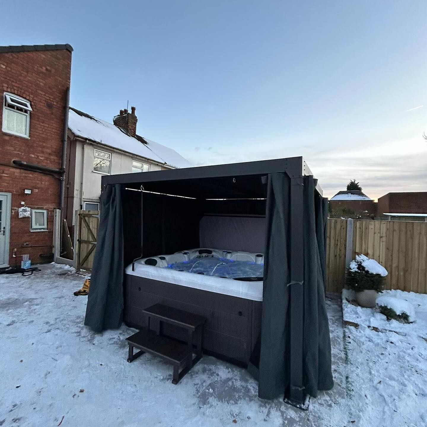 A hot tub covered by a dark canopy with drawn curtains. Snowy backyard scene with a brick building.