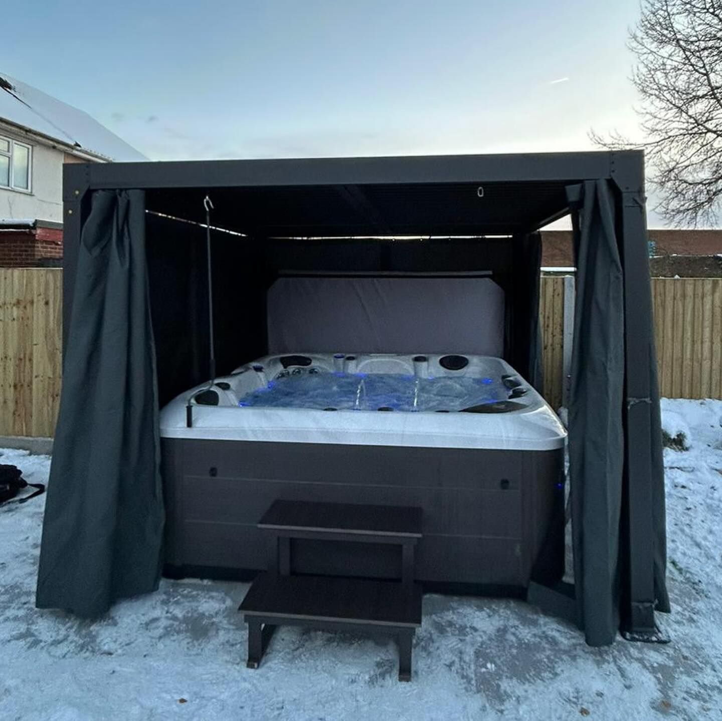 Hot tub enclosed by a dark canopy with drawn curtains; small step stool in front. Winter scene with snow.