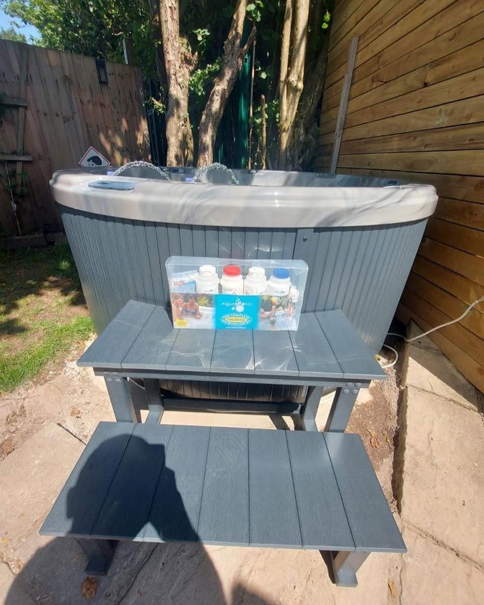 Hot tub with supplies on a gray two-tier step stool, set outdoors near a wooden fence and trees.
