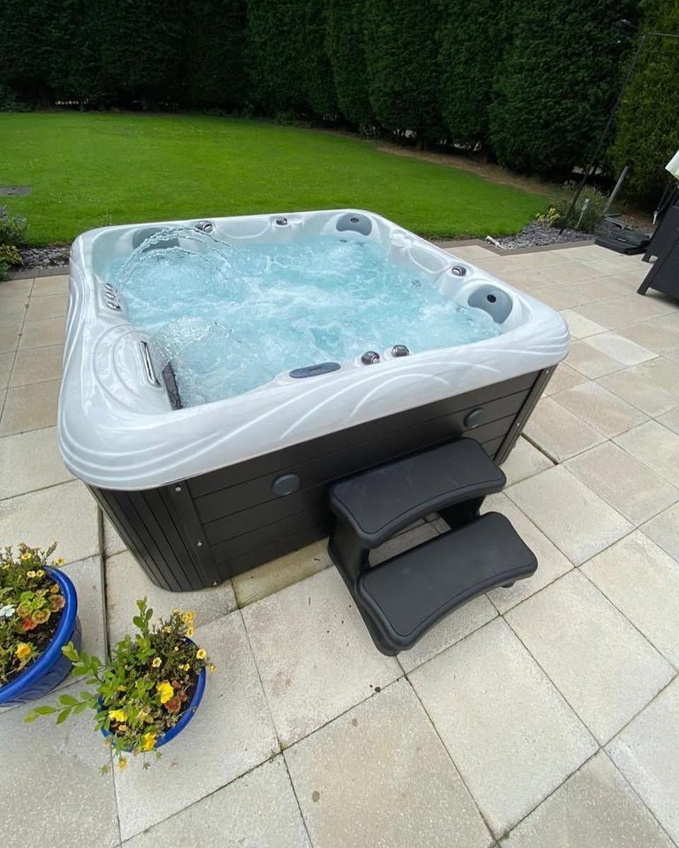 Hot tub on a patio, with steps, surrounded by green grass and plants. Water is bubbling.