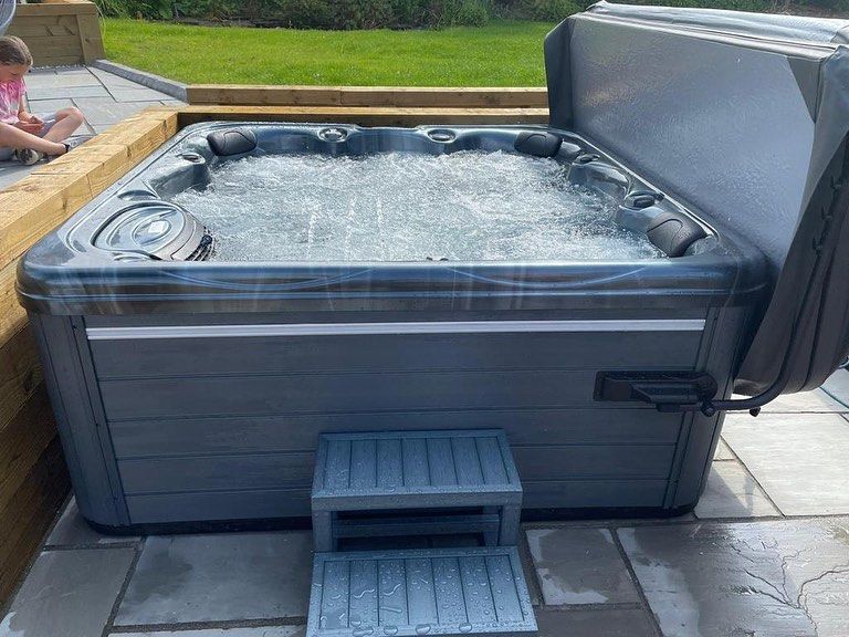 Bubbling hot tub with open cover, gray exterior, and step stool. Set outdoors on patio.