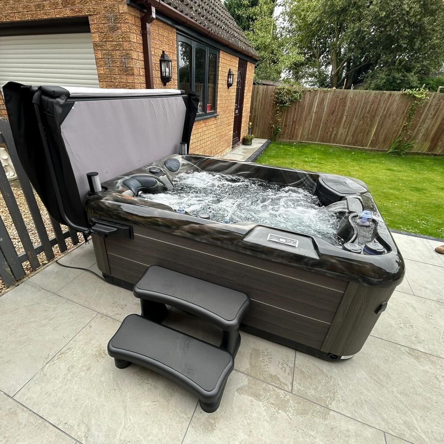 Hot tub on a patio with steps. The cover is open, water is bubbling. The setting is outdoors.