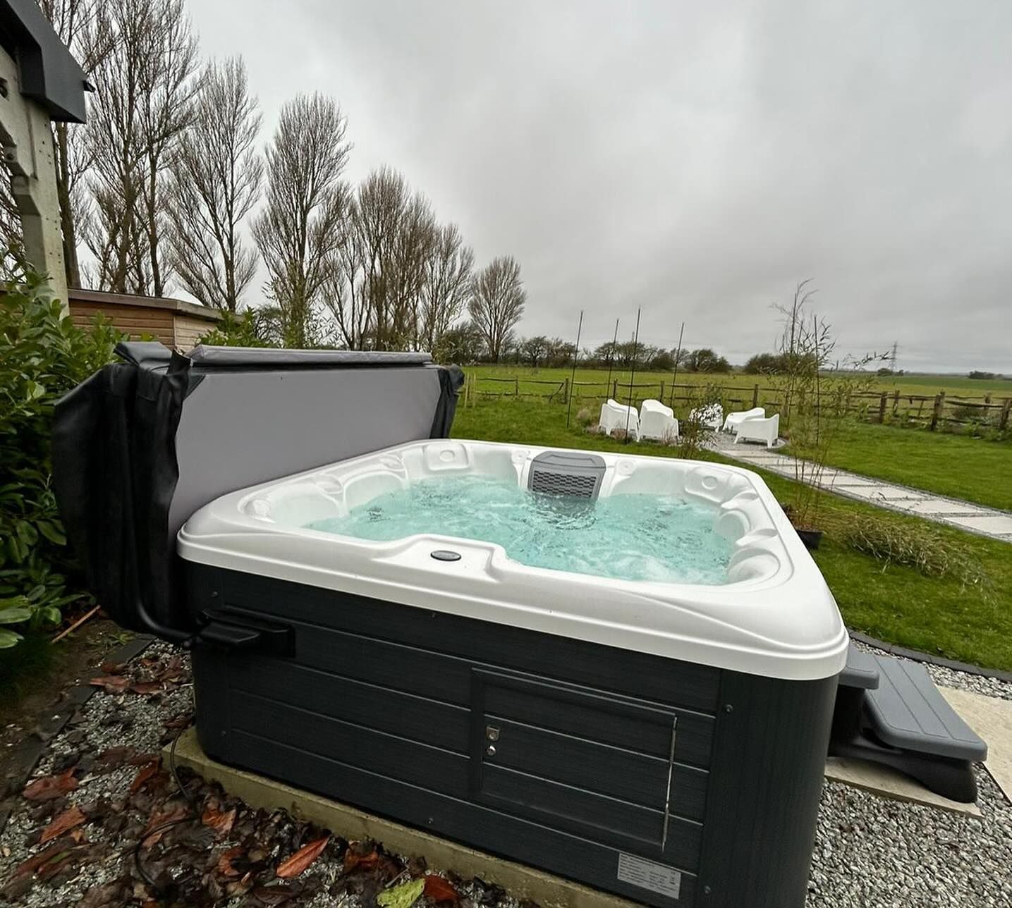 Hot tub outdoors, gray cover open, bubbling water. Gray and black spa, overcast day, grassy field background.