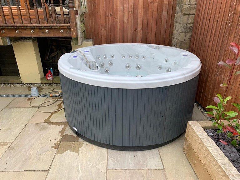 Round hot tub with gray exterior, set on a stone patio beside a wooden fence and planter.