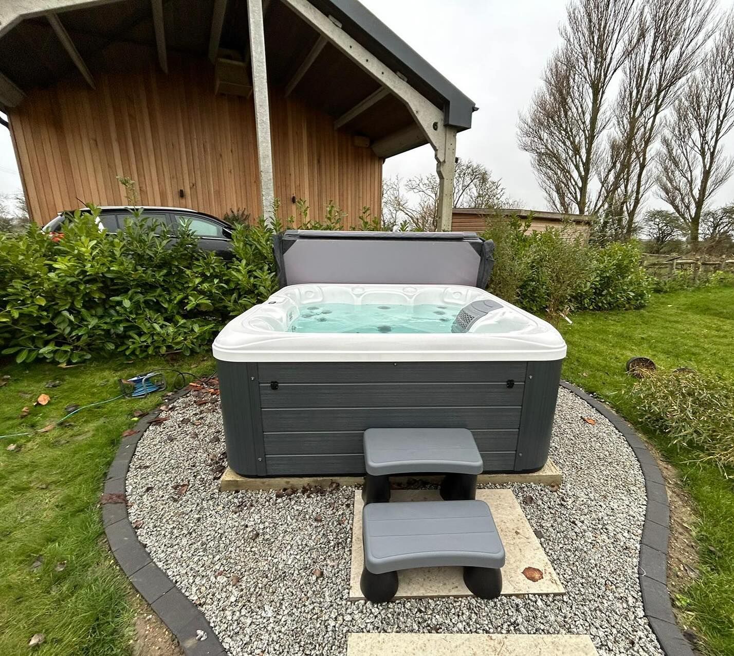 Hot tub on a stone base, with steps in front. Grey tub, light water, and outdoor setting.