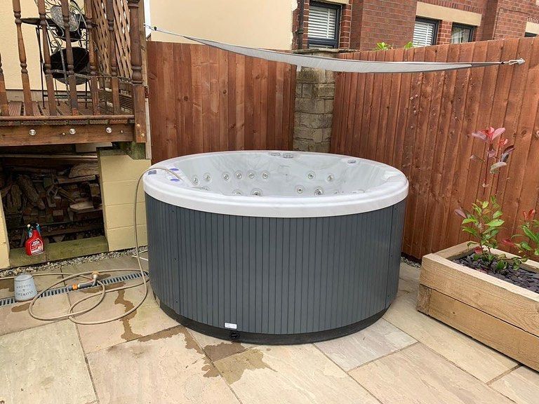 Round hot tub, grey and white, on stone patio, near wooden fence and planter.