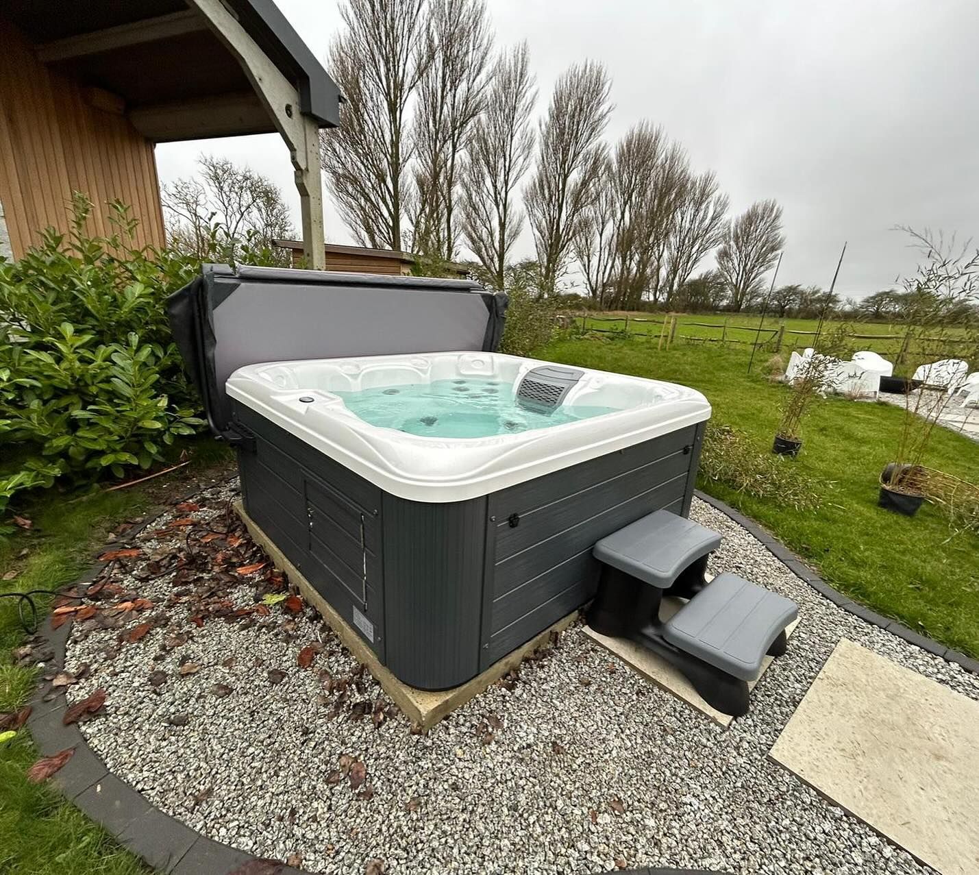 Hot tub, dark grey exterior, partially open cover, two-step grey stairs. Outdoors, gravel base, green lawn, cloudy sky.