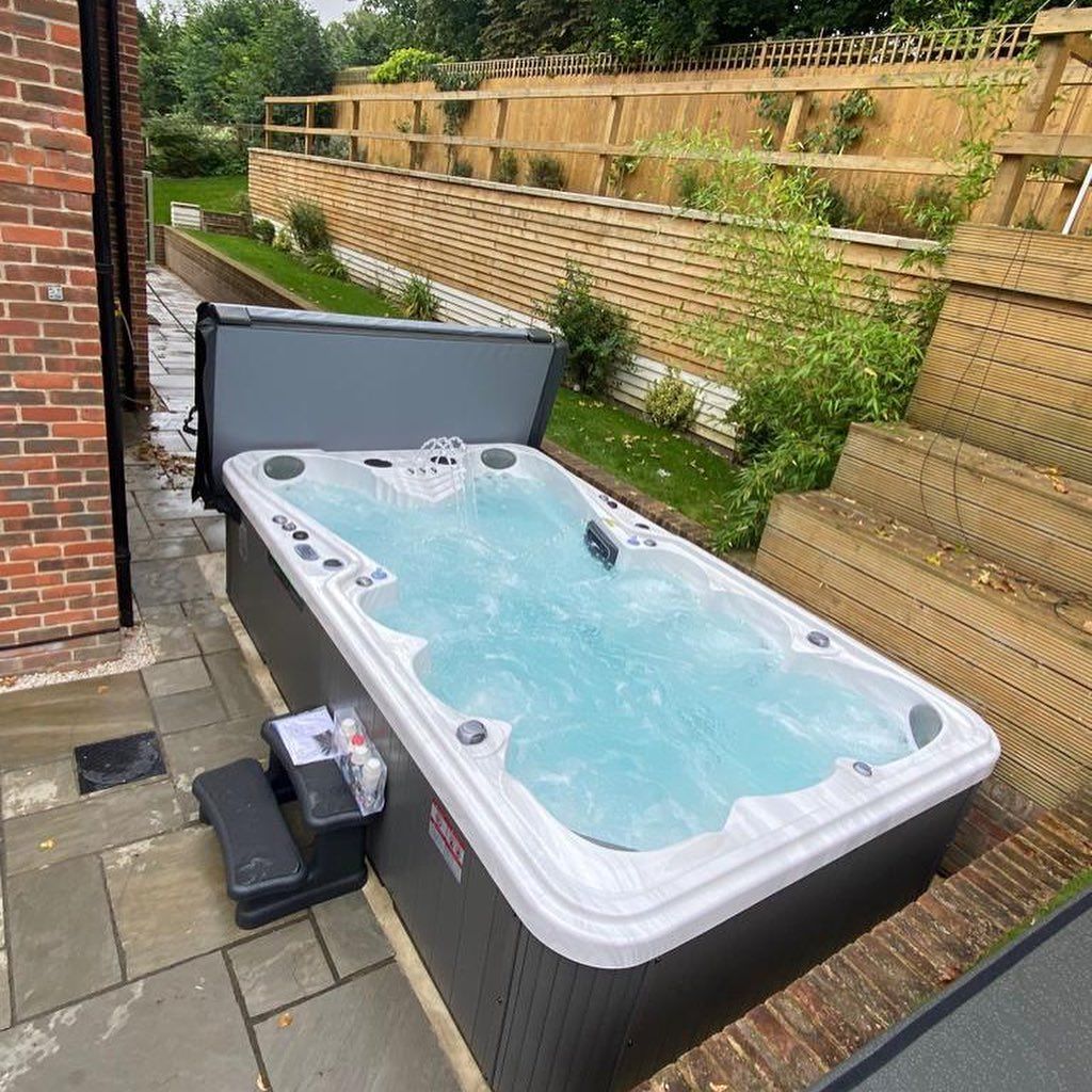 Rectangular swim spa with open cover on patio near a terraced garden with wooden fence.