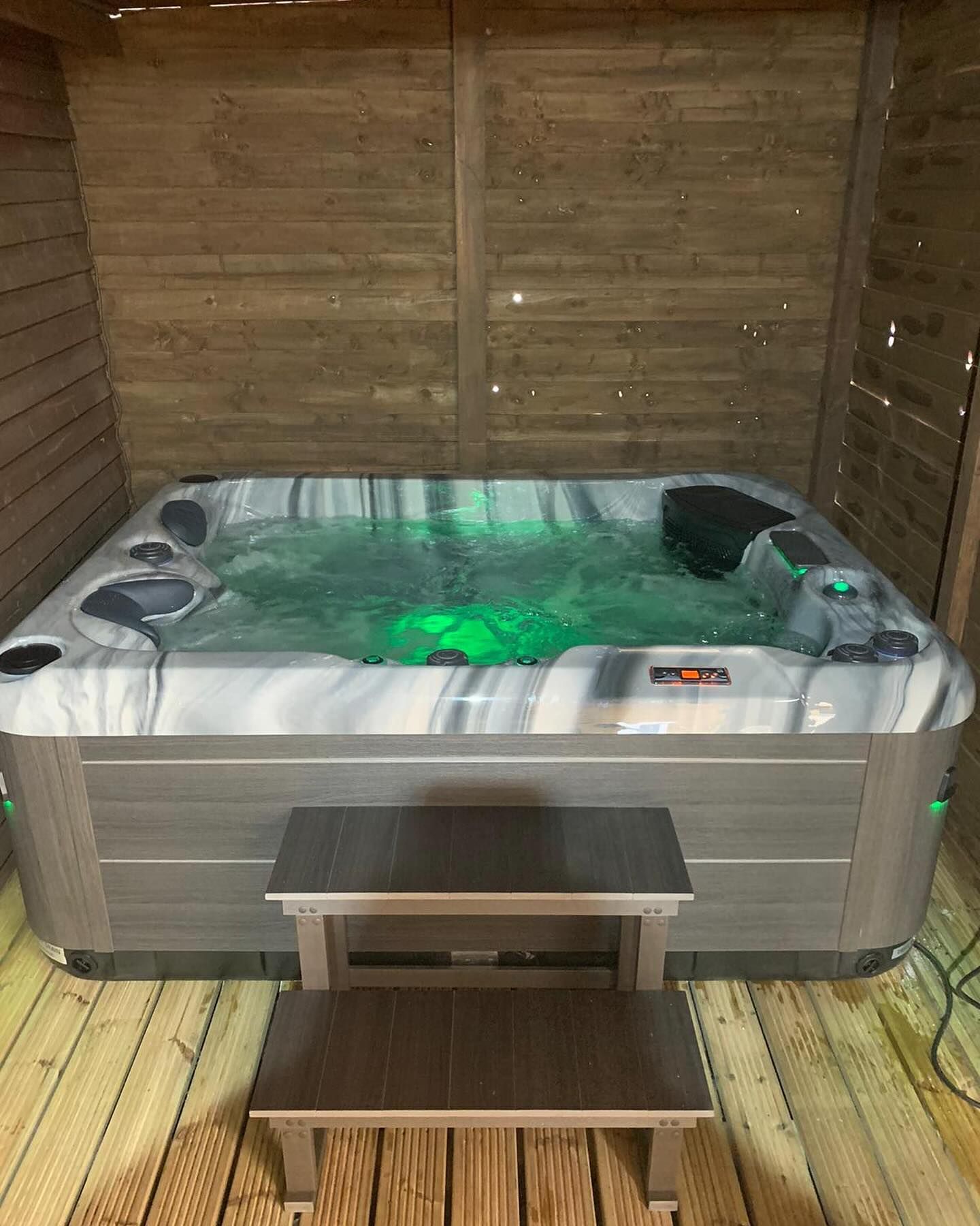 Hot tub with steps on a wooden deck inside a wooden enclosure; green underwater lights.