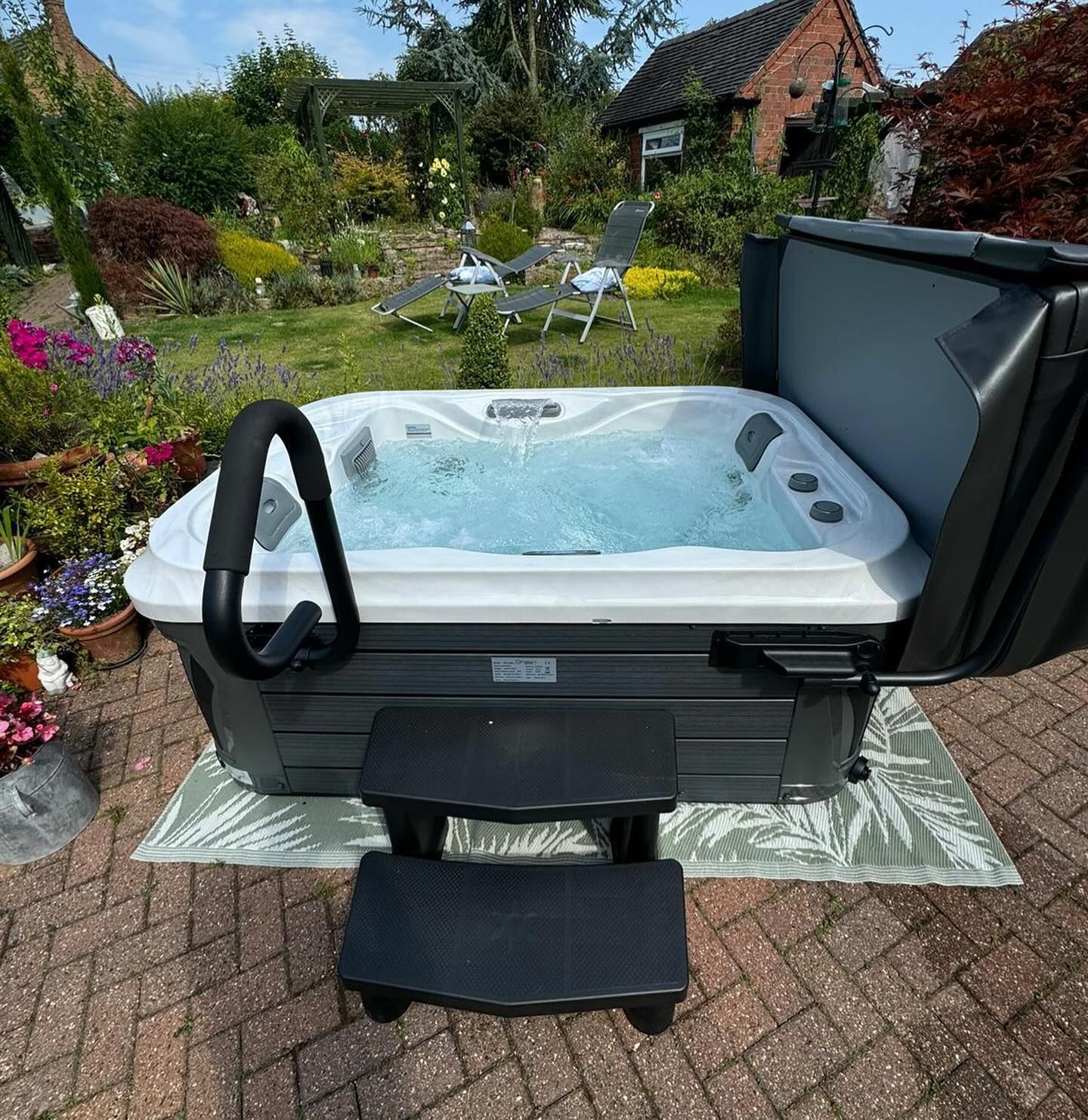 Hot tub in a backyard with a handrail, steps, and cover open. Green foliage and seating in background.