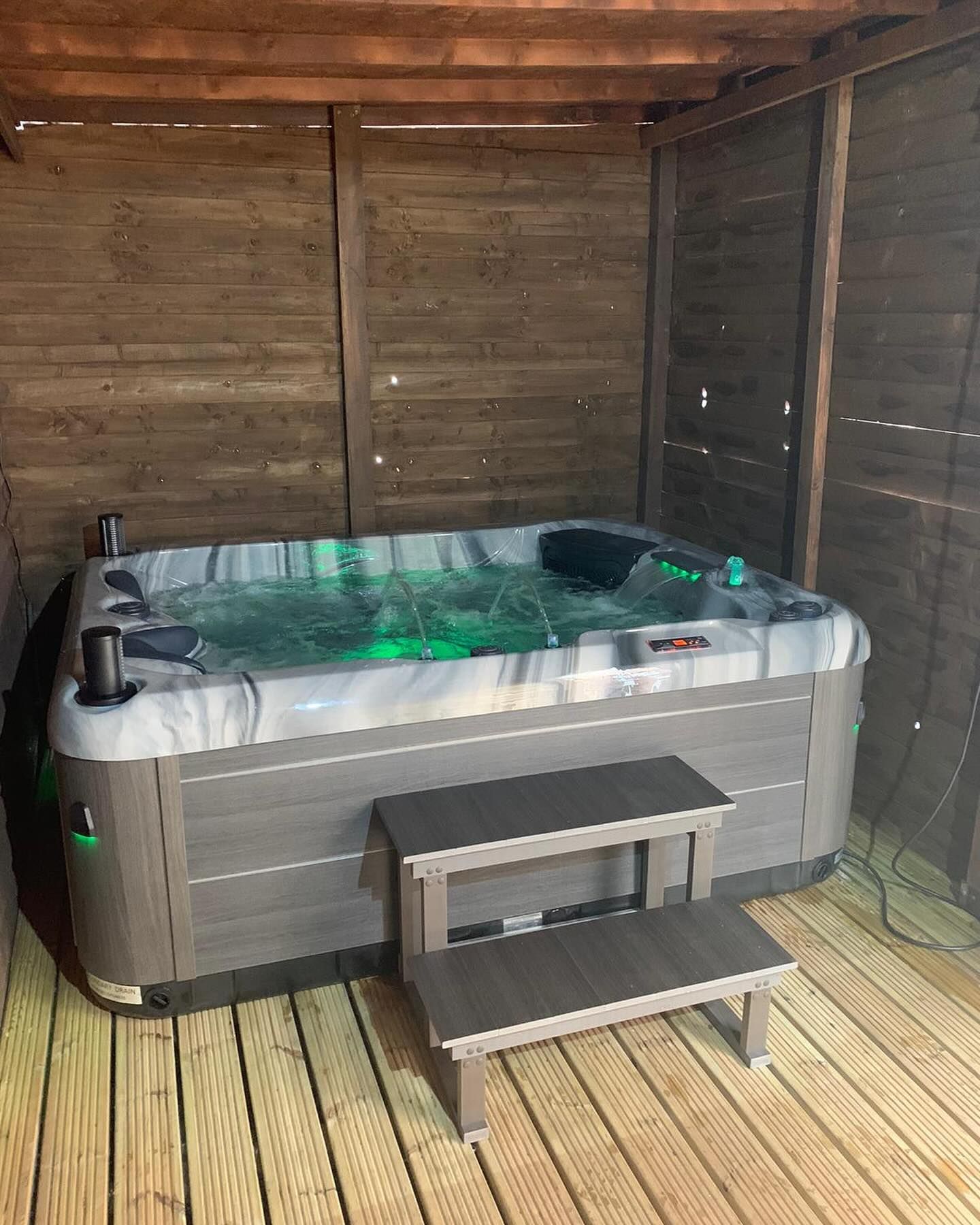 Hot tub with green glowing water, next to wooden walls and stairs.