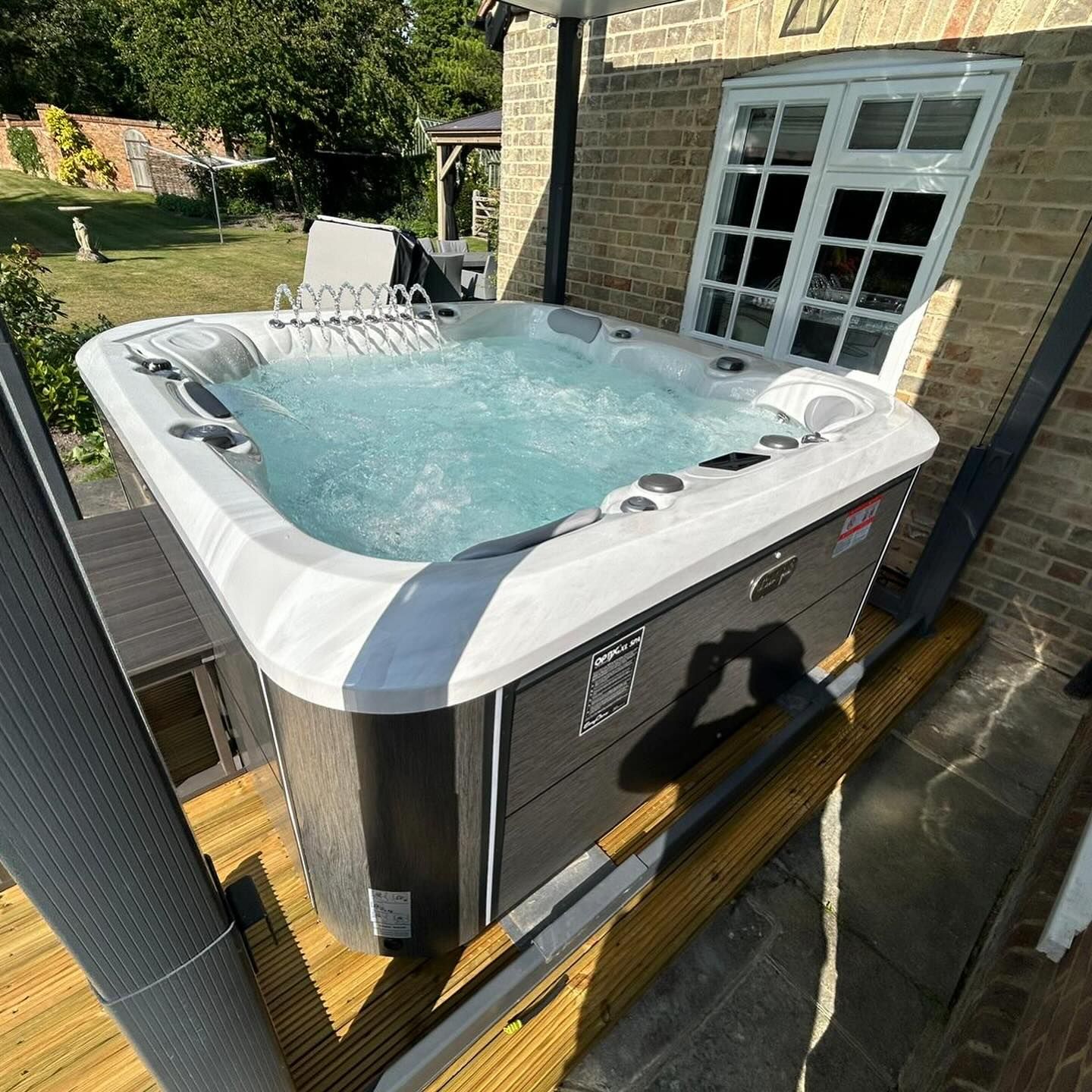 Hot tub on wooden deck, bubbling water, next to a brick building with white window.