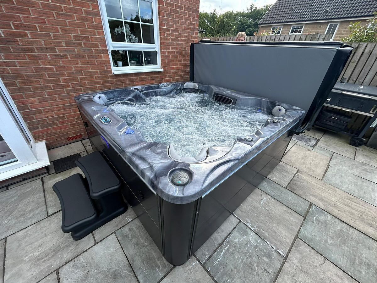 Hot tub with bubbling water on a paved patio, beside a brick wall and window. Grey cover is open.