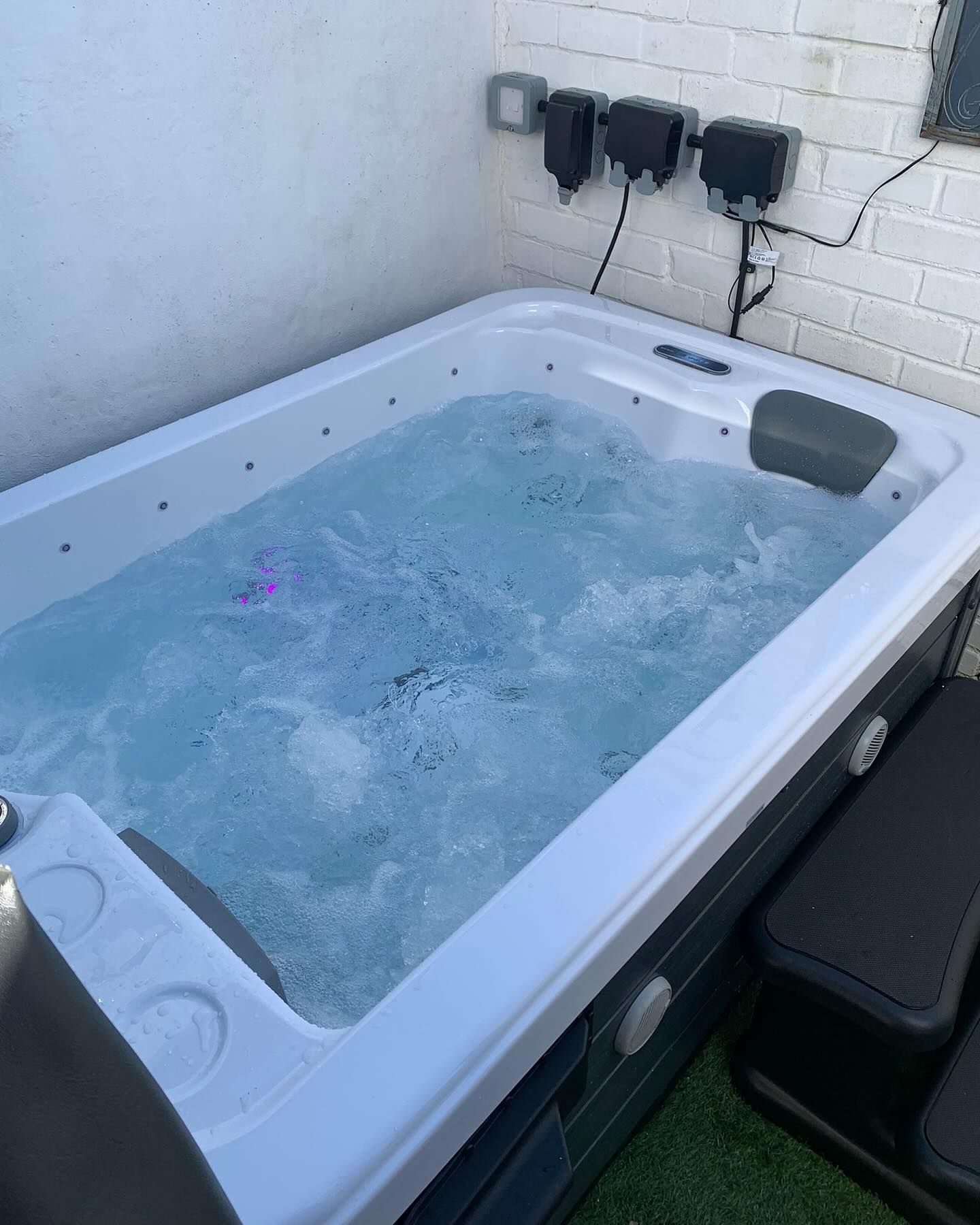 Rectangular hot tub filled with bubbling water, against a white brick wall, and black control boxes above.