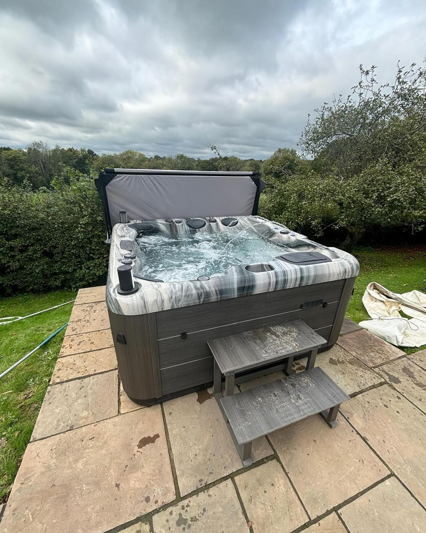 Hot tub with cover open, on a patio. Gray and black exterior with wooden steps. Lush green trees in background. Cloudy sky.