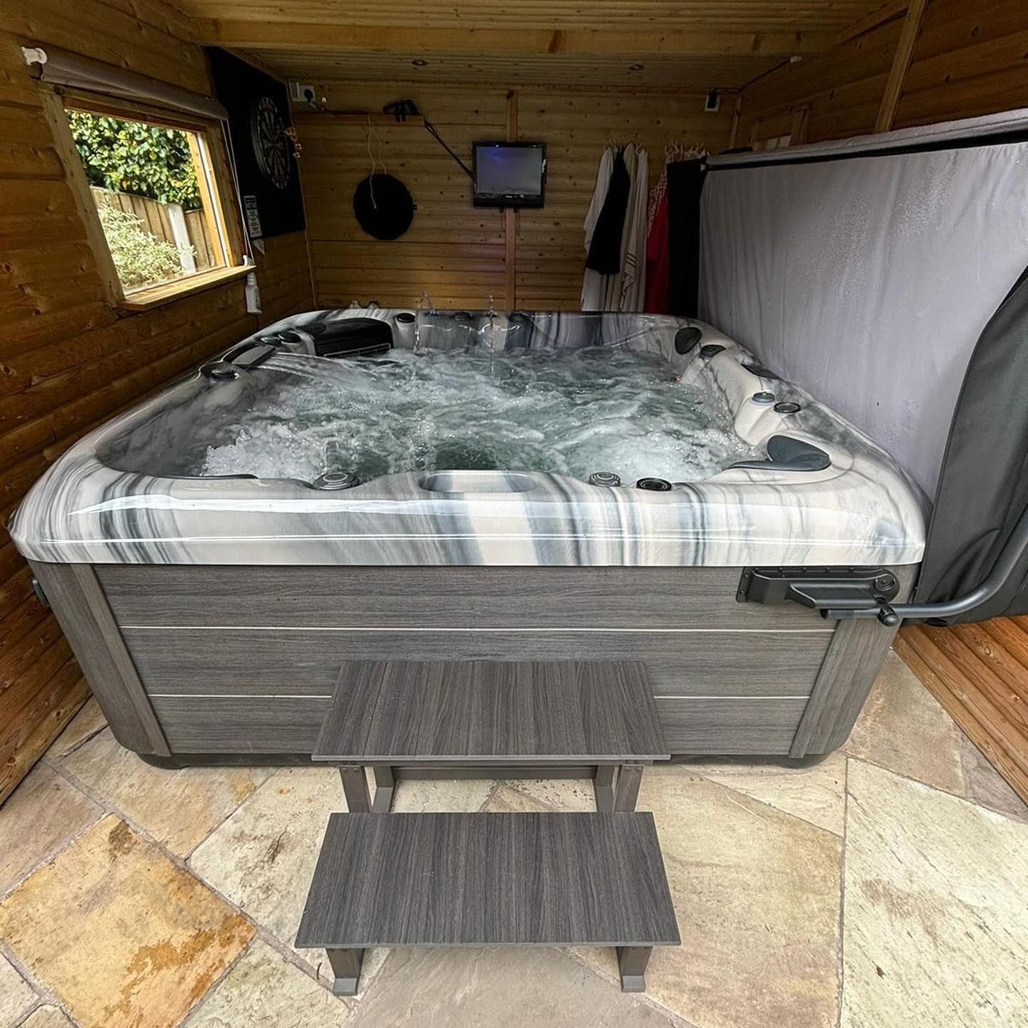 Hot tub in a wooden enclosure with steps. Water is bubbling, and a small TV is mounted on the wall.