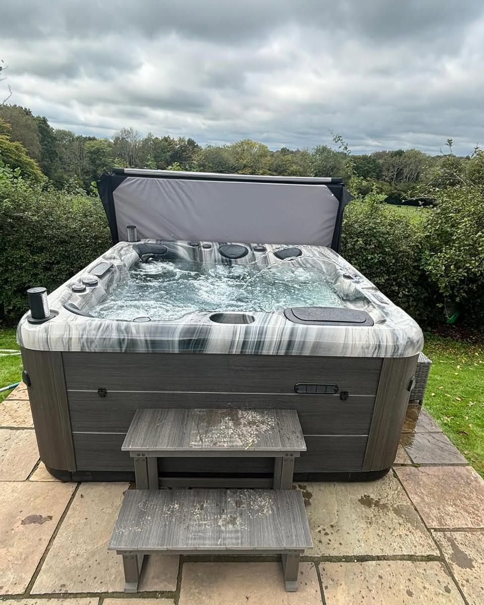 Hot tub on a stone patio with steps; grey water, cover, and siding; overcast sky.