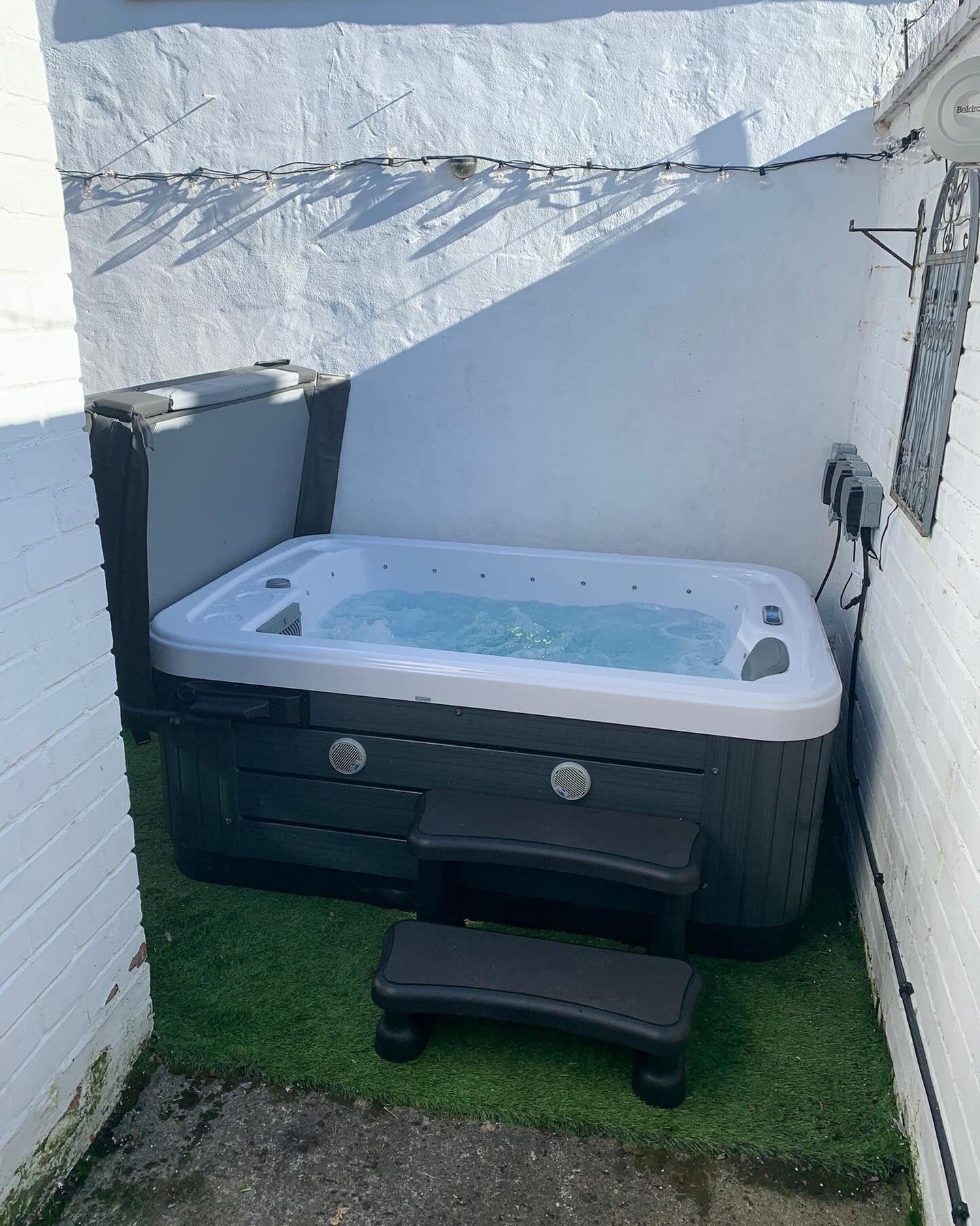 Hot tub in a small outdoor area, dark gray with steps and cover, against white brick walls, artificial green grass.