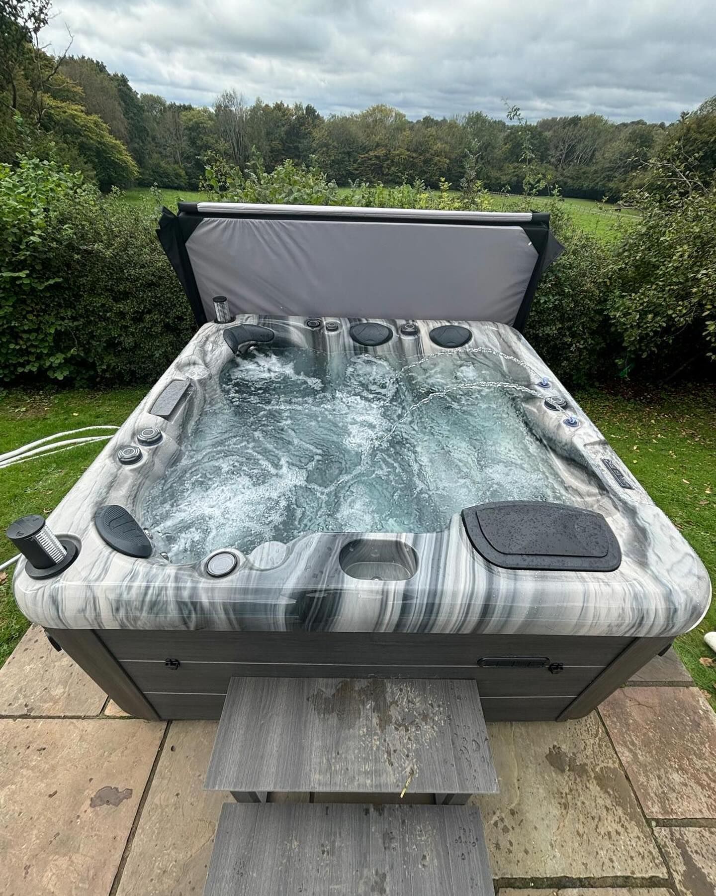 Hot tub on a stone patio with cover open; water bubbling. Green lawn and trees in background.