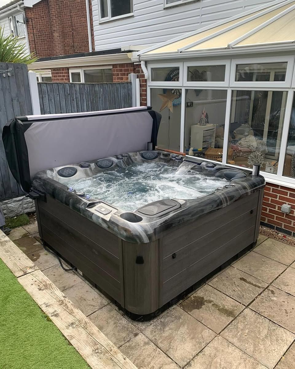 Hot tub with open cover on a patio next to a building with windows. Gray and brown exterior.