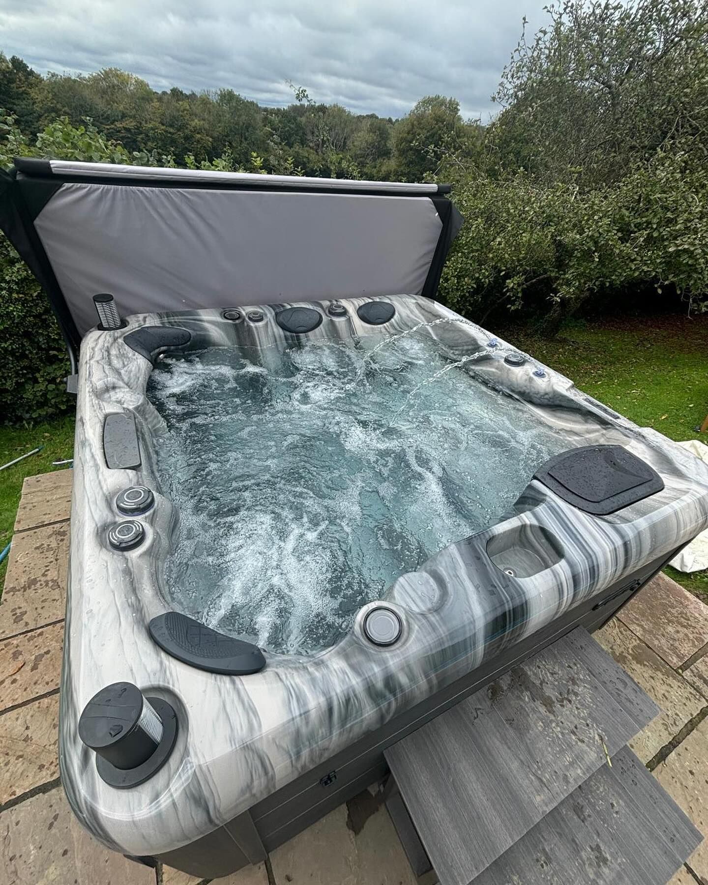 A hot tub with swirling water, partially covered, on a stone patio with greenery in the background.