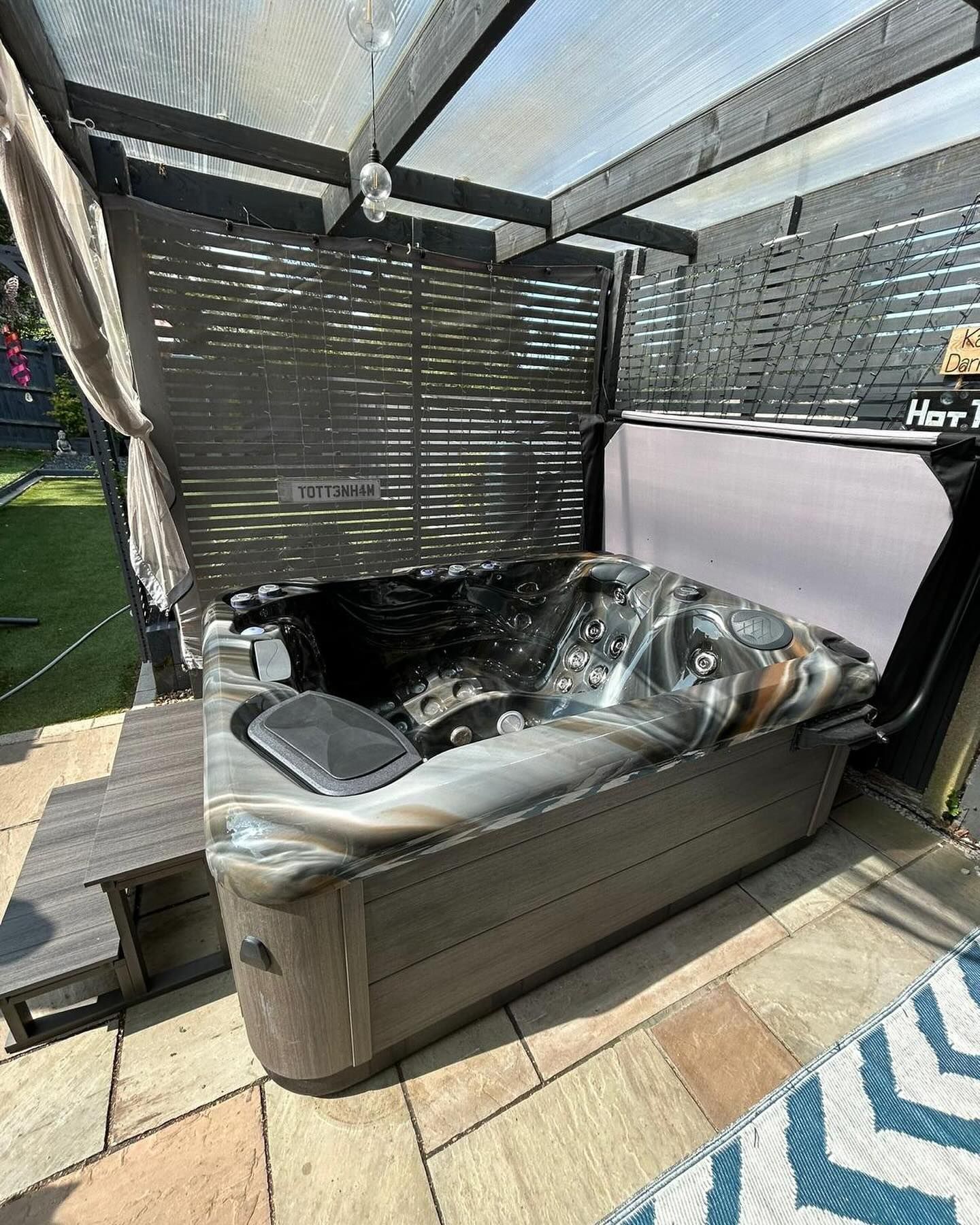 Hot tub on patio under a black pergola. Gray tub with open cover, wooden bench, and gray privacy screen.