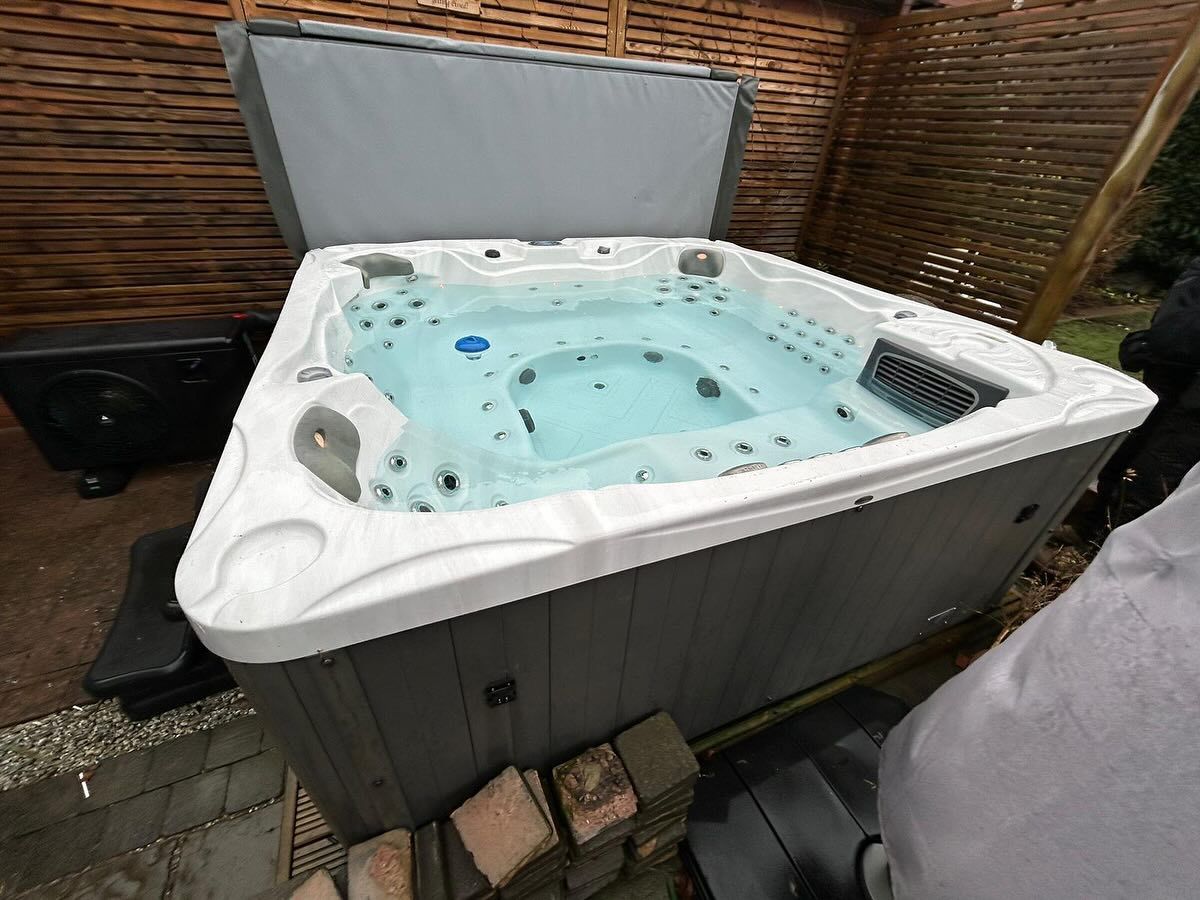 Hot tub with open cover, filled with water, placed outdoors. Grey and white design with surrounding wooden fence.