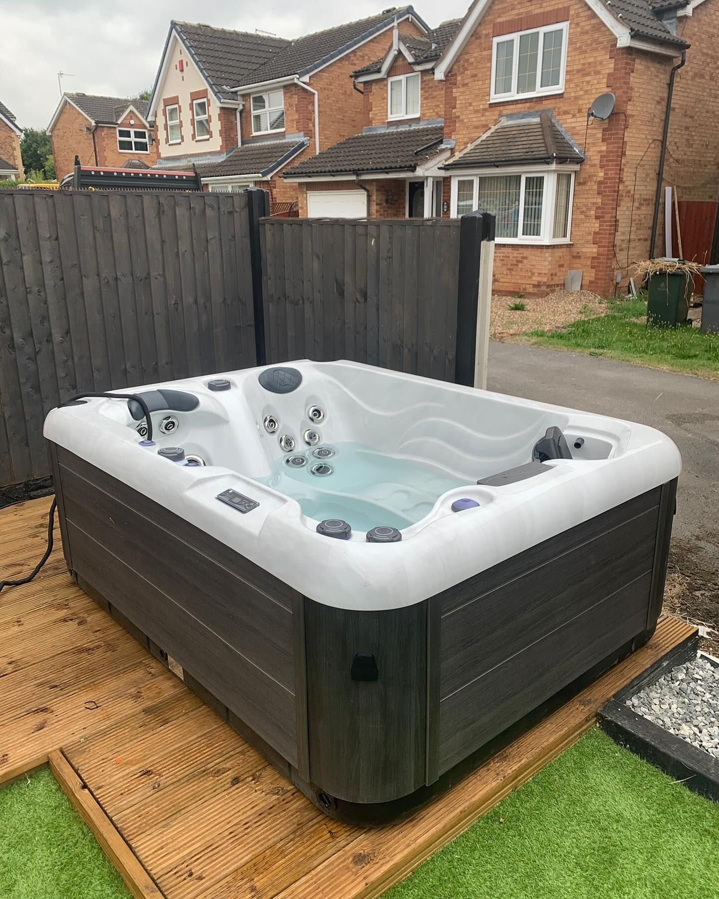 Hot tub on wooden deck in a backyard with a gray fence and brick houses in the background.