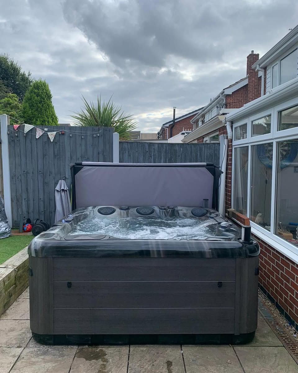 Hot tub in a backyard, grey cover partially open. Cloudy sky, gray fence, and brick house.