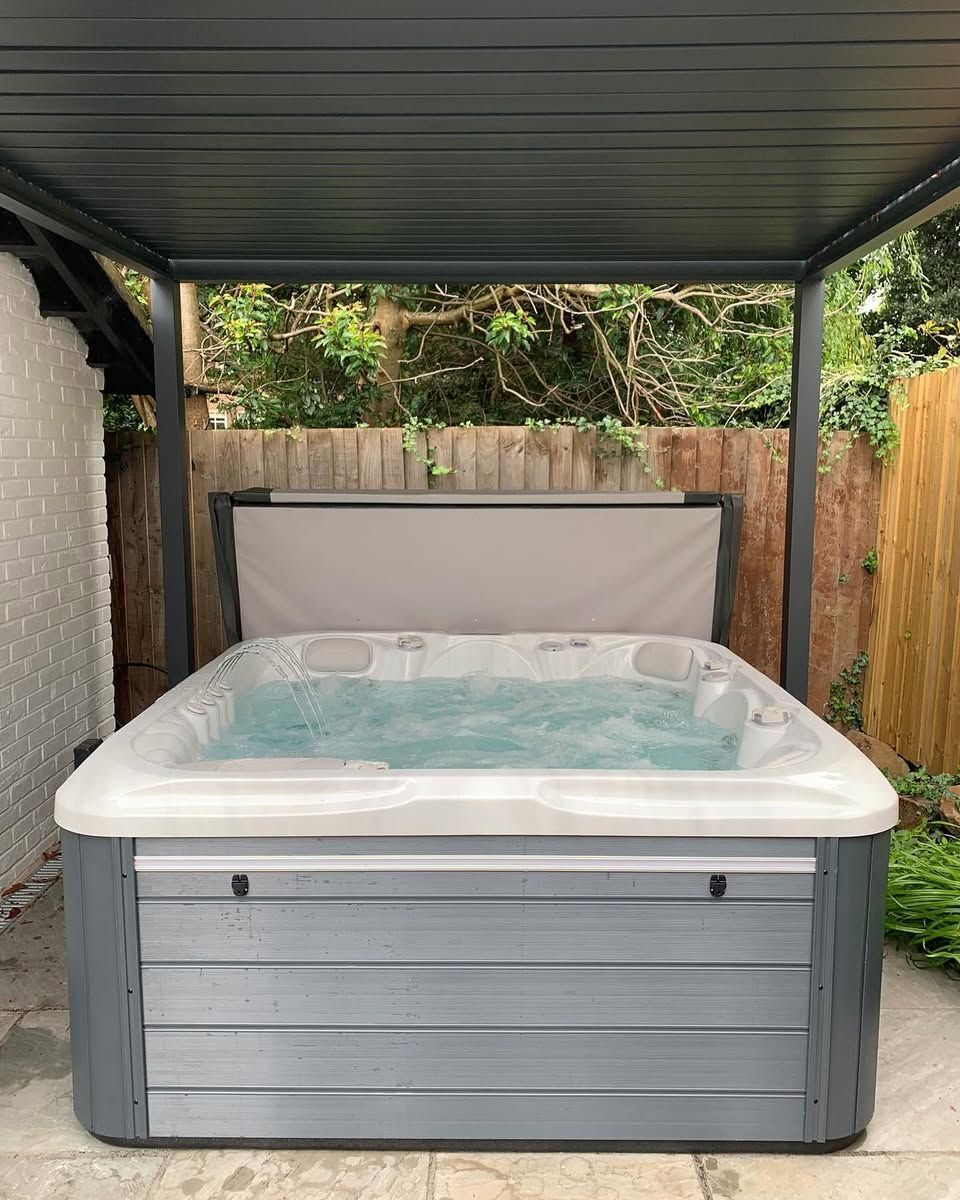 Hot tub filled with water, under a gray gazebo. Gray siding, surrounded by a wooden fence and greenery.
