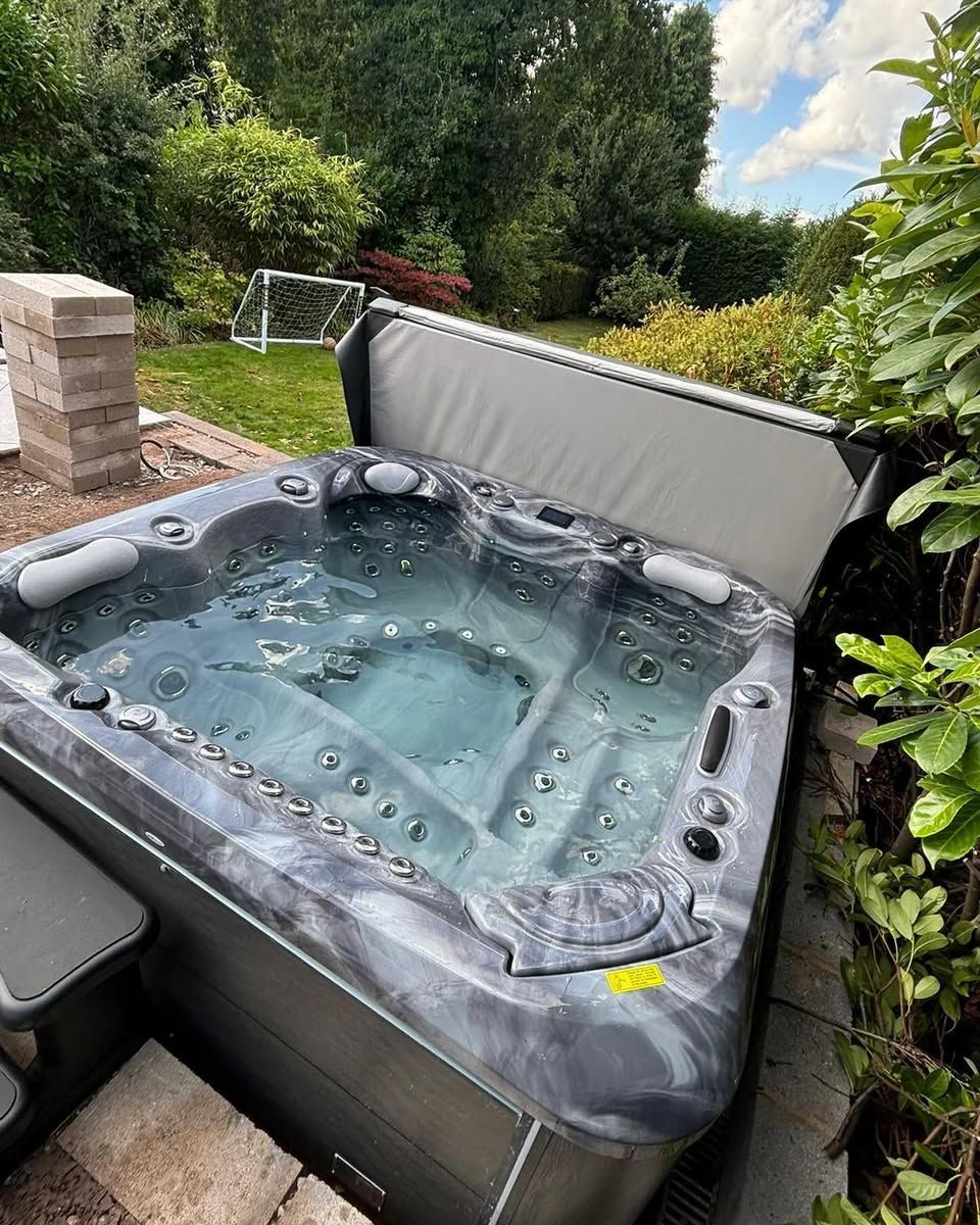 Hot tub filled with water in a backyard setting. Cover partially open, surrounded by greenery.