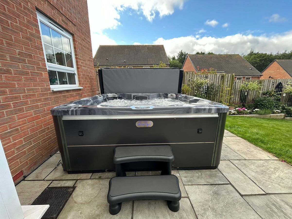 Black hot tub with gray steps on a stone patio next to a brick building.