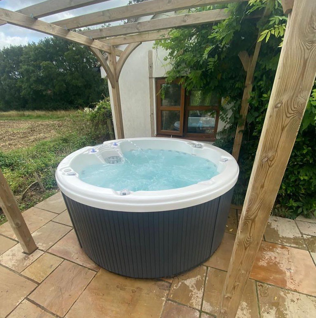 Round hot tub with bubbling water sits under a wooden pergola on a stone patio outdoors.
