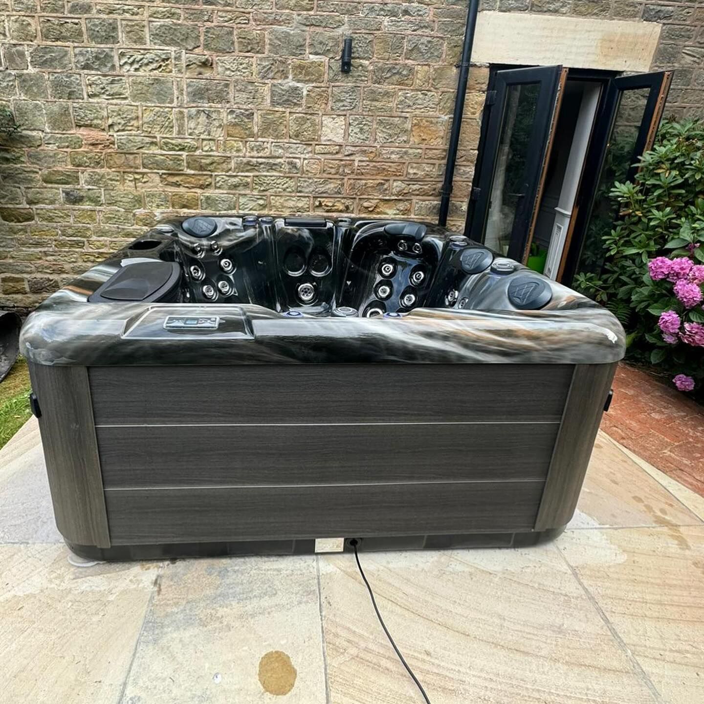 Hot tub with dark gray and brown exterior, set on stone patio in front of a stone wall.