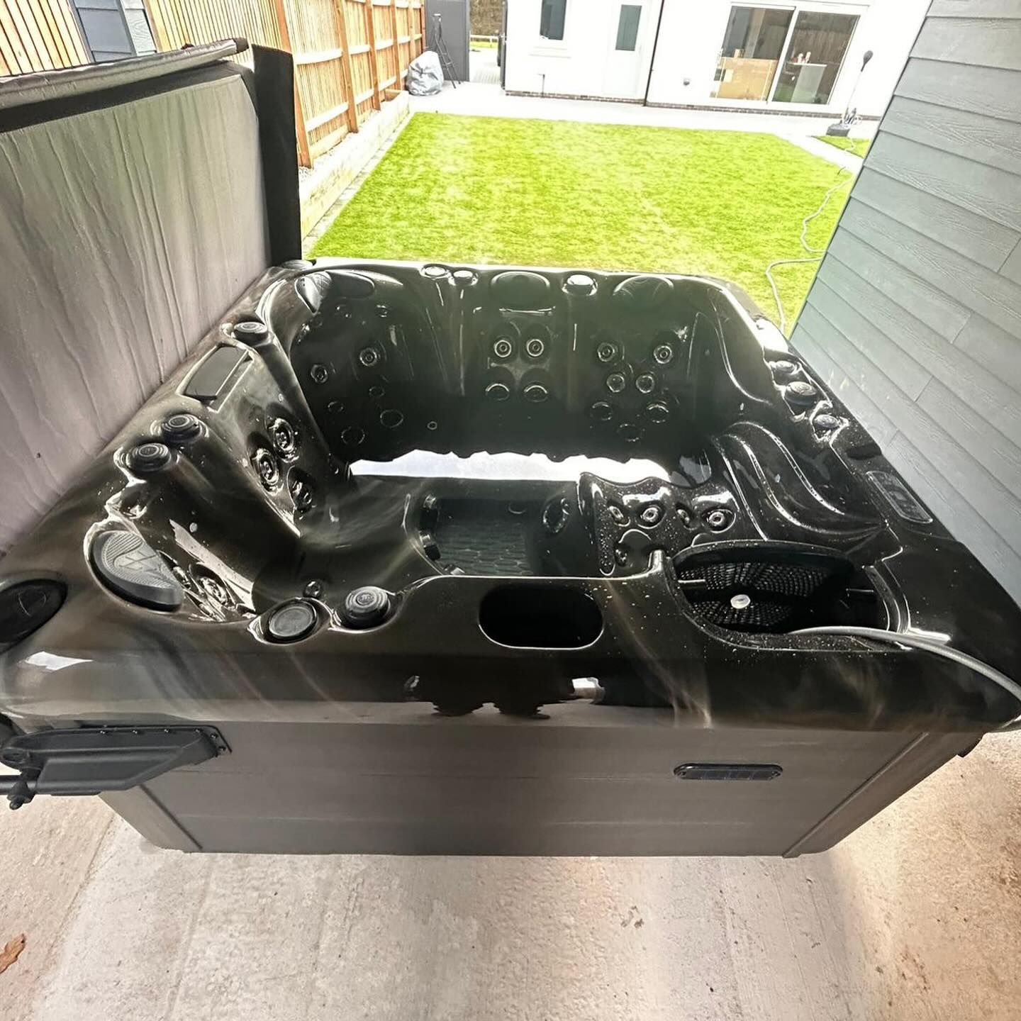 Black hot tub with gray cabinet, open lid, on patio with green lawn background.