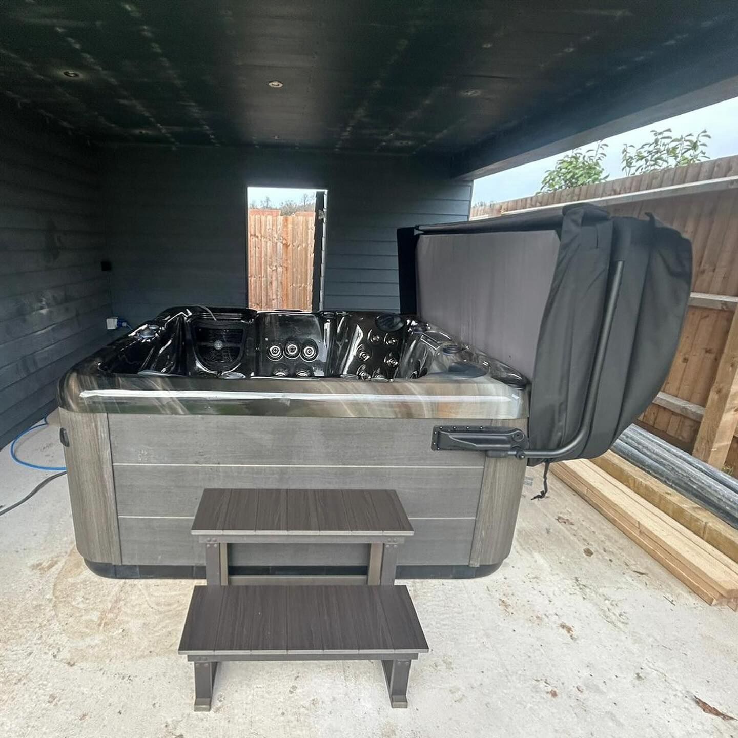 Hot tub with open cover and steps, under a dark gray shelter.