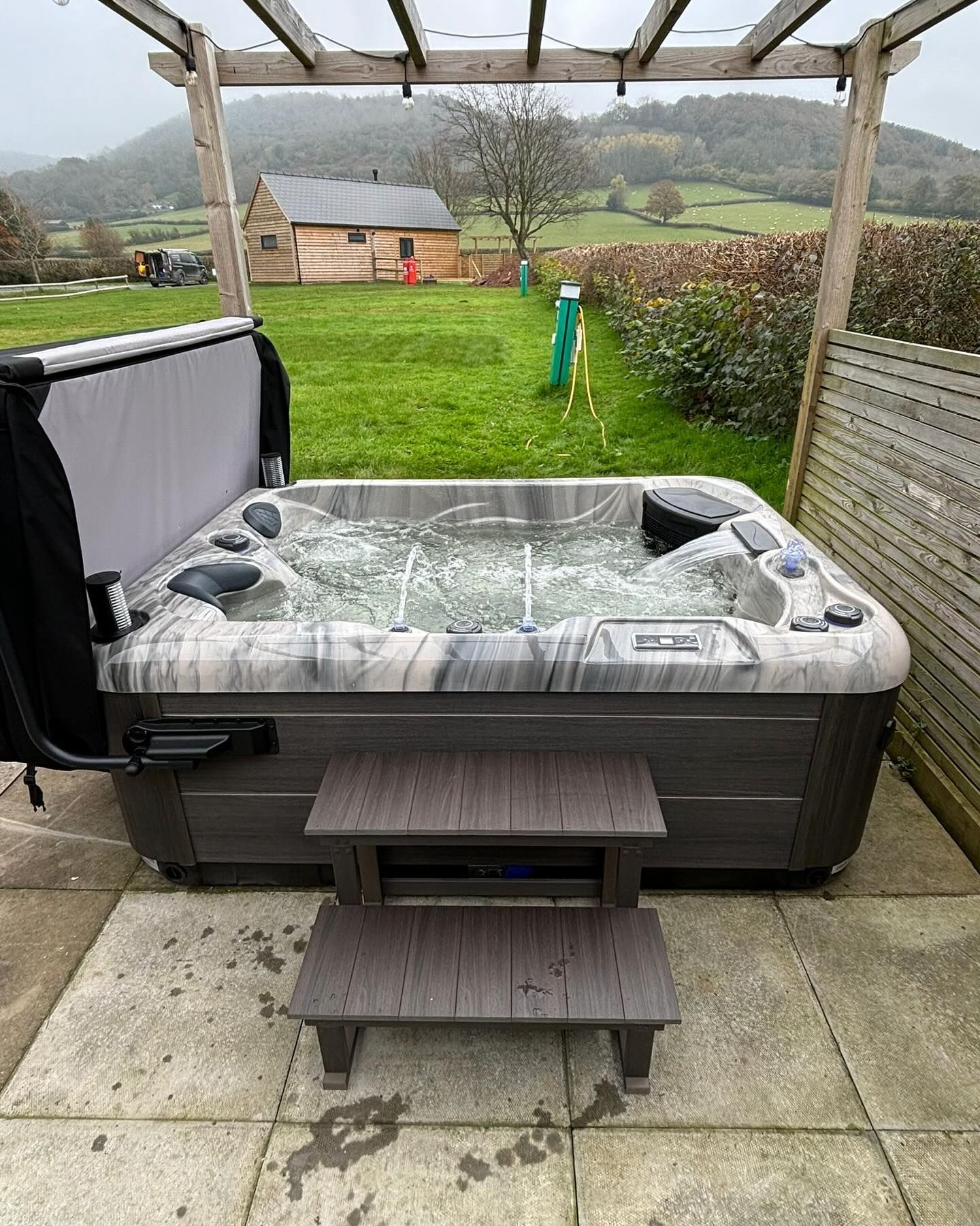 Hot tub with steps, outdoors. Gray and white tub with water and jets. Wooden pergola overhead, green grass and cloudy sky.