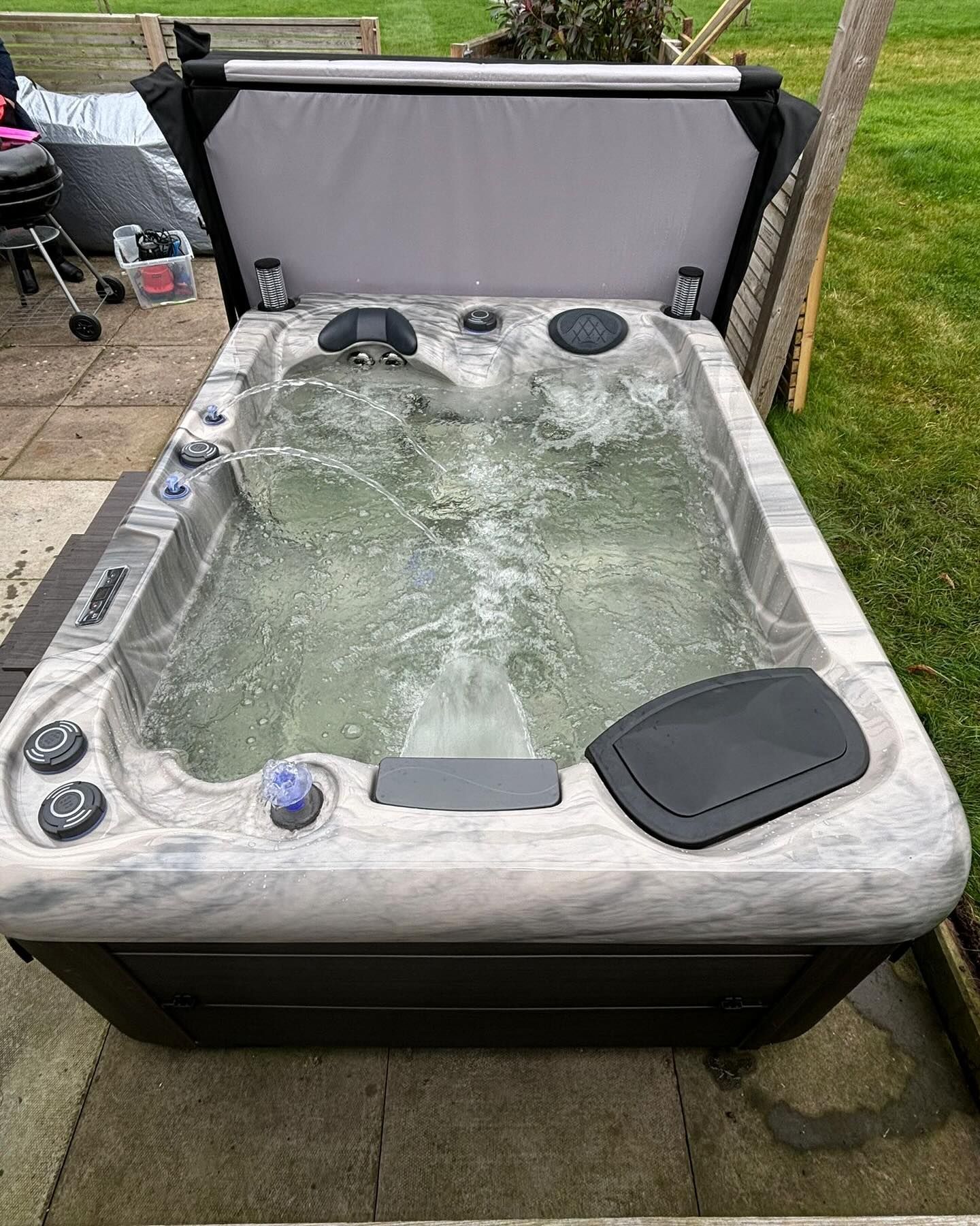 Hot tub filled with water, grey and black colors, partially covered, outdoors on a patio.