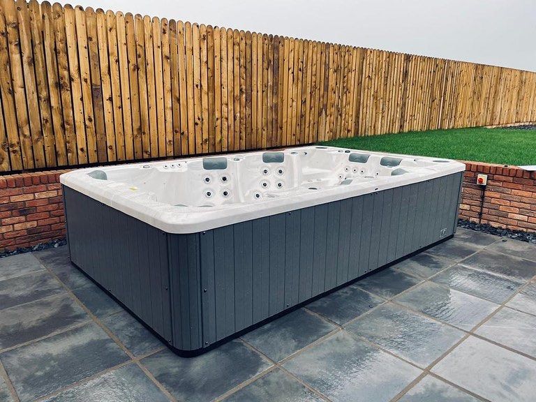 Rectangular gray hot tub on a tiled patio, with a wooden fence and green grass in the background.
