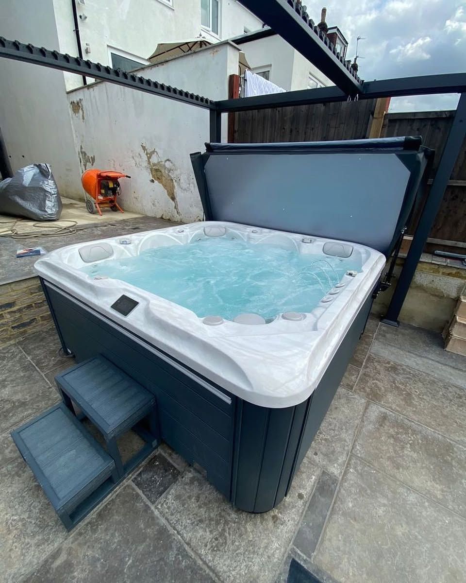 Hot tub with open cover, steps, and bubbling water, set in a backyard. Blue-gray exterior with a dark pergola.