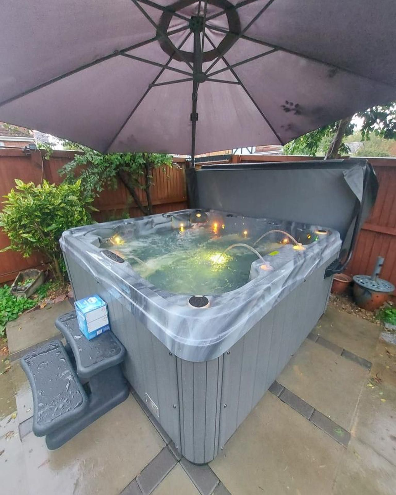 Hot tub under a gray umbrella in a backyard, water bubbling. Steps lead up to it, cover is partially open.