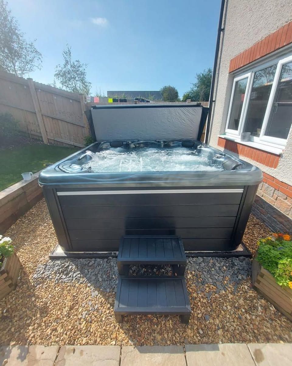 Hot tub with open cover, two-step entry, on gravel, beside a house with a window, on a sunny day.
